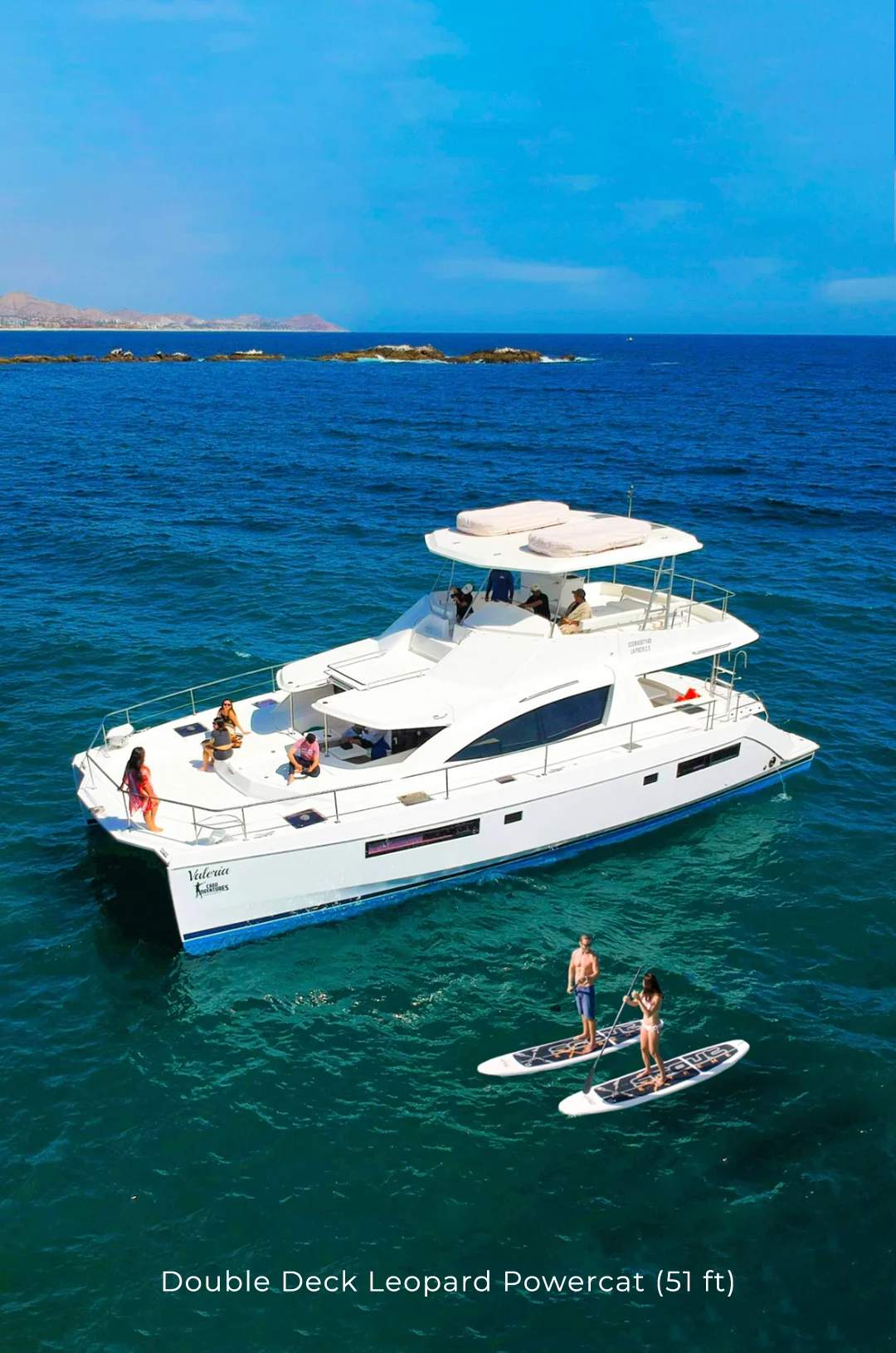 Yacht anchored with people paddleboarding and swimming in clear blue waters near the coast.