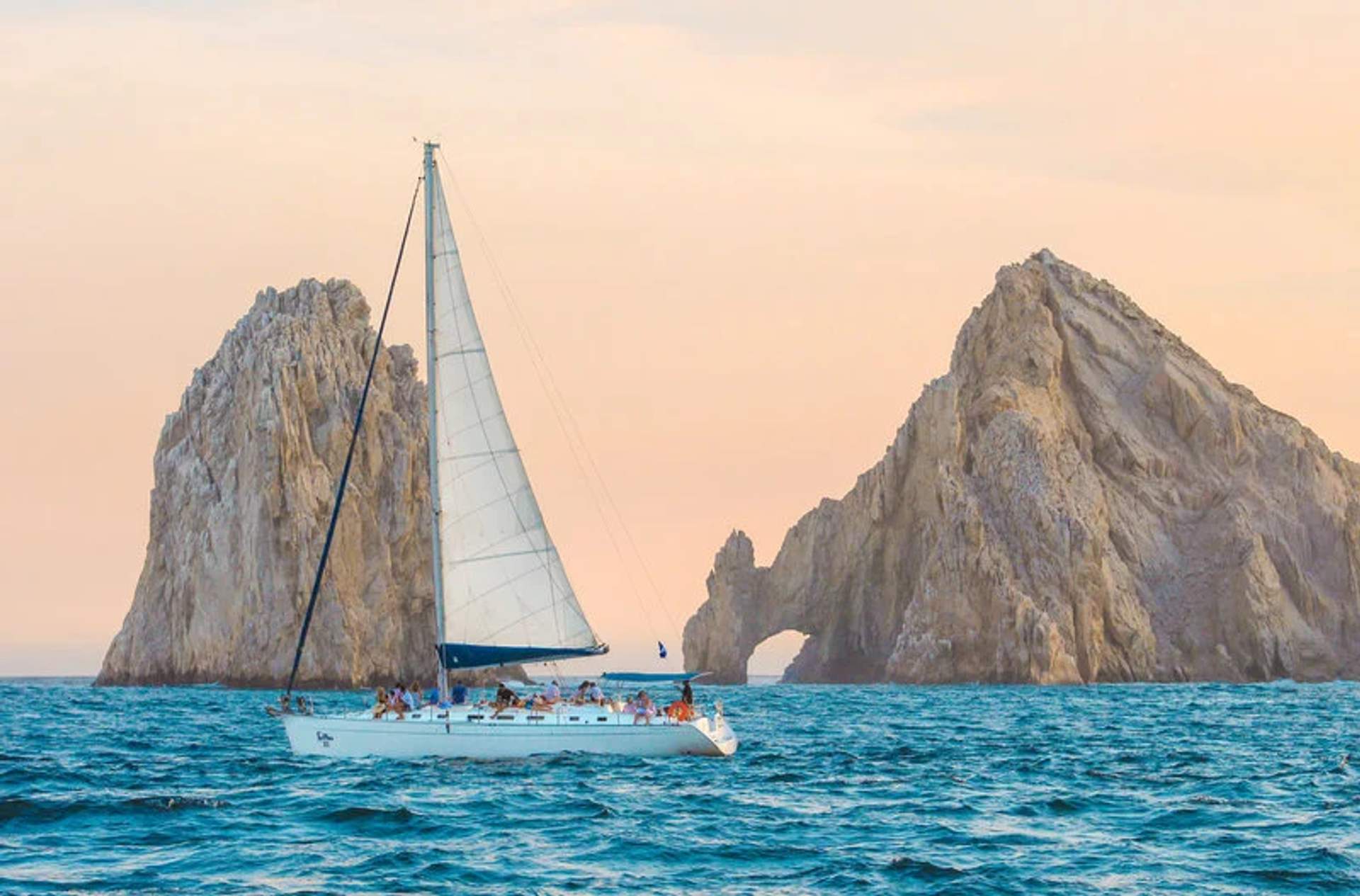 A sailboat with several passengers sails on a calm ocean with rocky formations in the background under a pastel-colored sky.