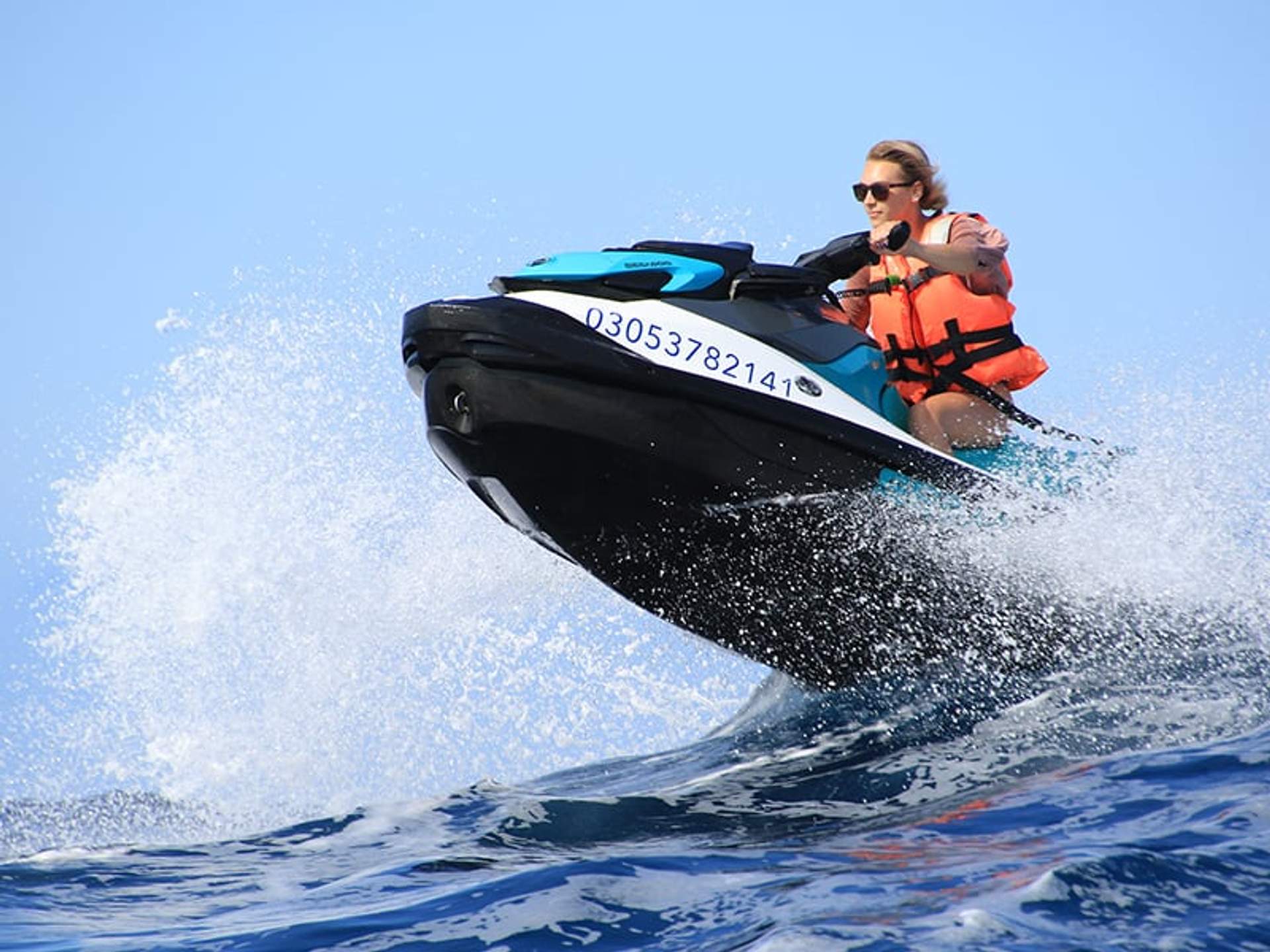 A woman wearing a life vest rides a jet ski, catching air over a wave on a sunny day.