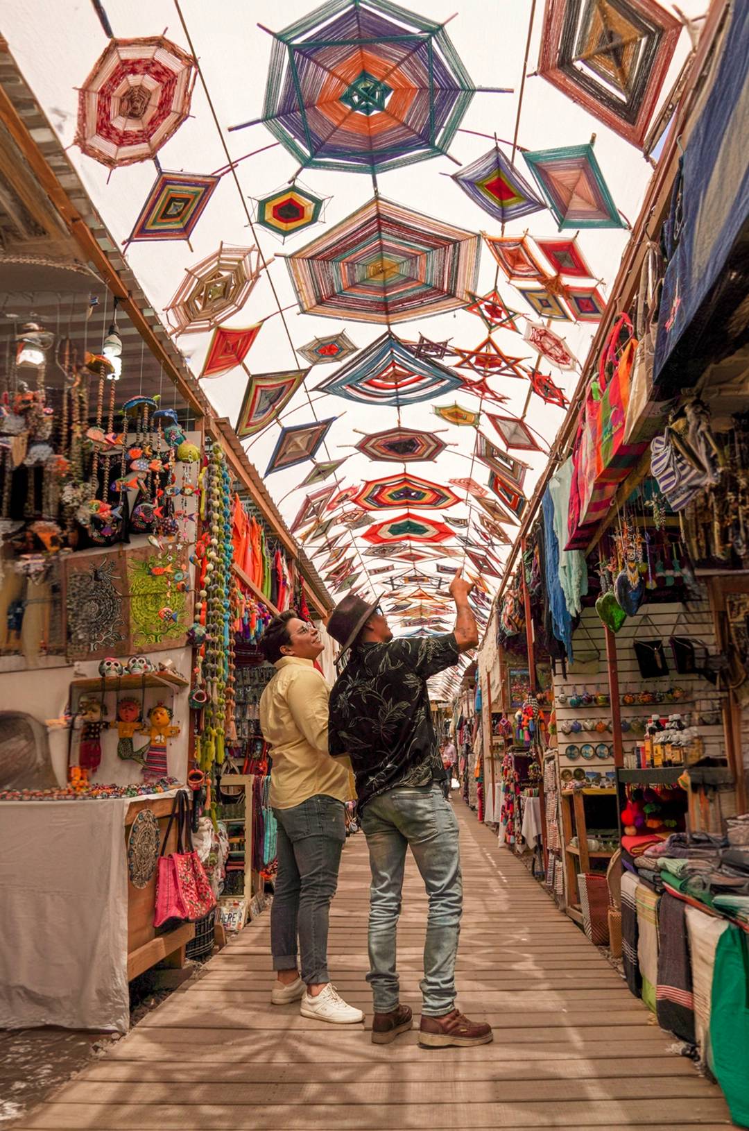 Two people in a market looking at colorful crafts and decorations, with vibrant woven designs hanging from the ceiling.
