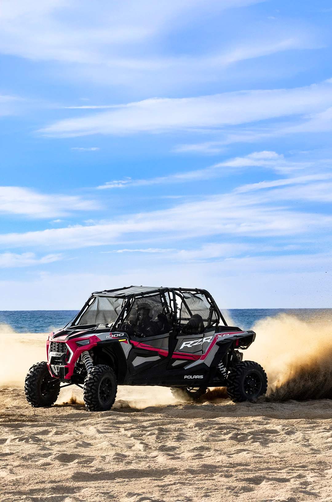 A UTV kicks up sand as it speeds along a beach with the ocean in the background, under a partly cloudy sky.