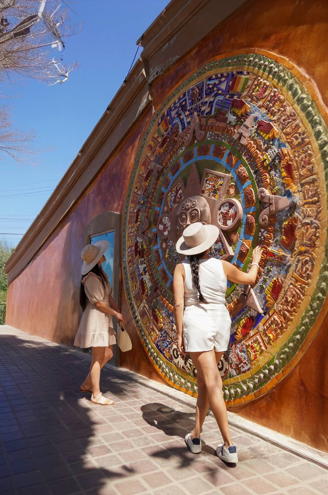 Two women in hats admiring a large, colorful Aztec calendar mural on a sunny day.