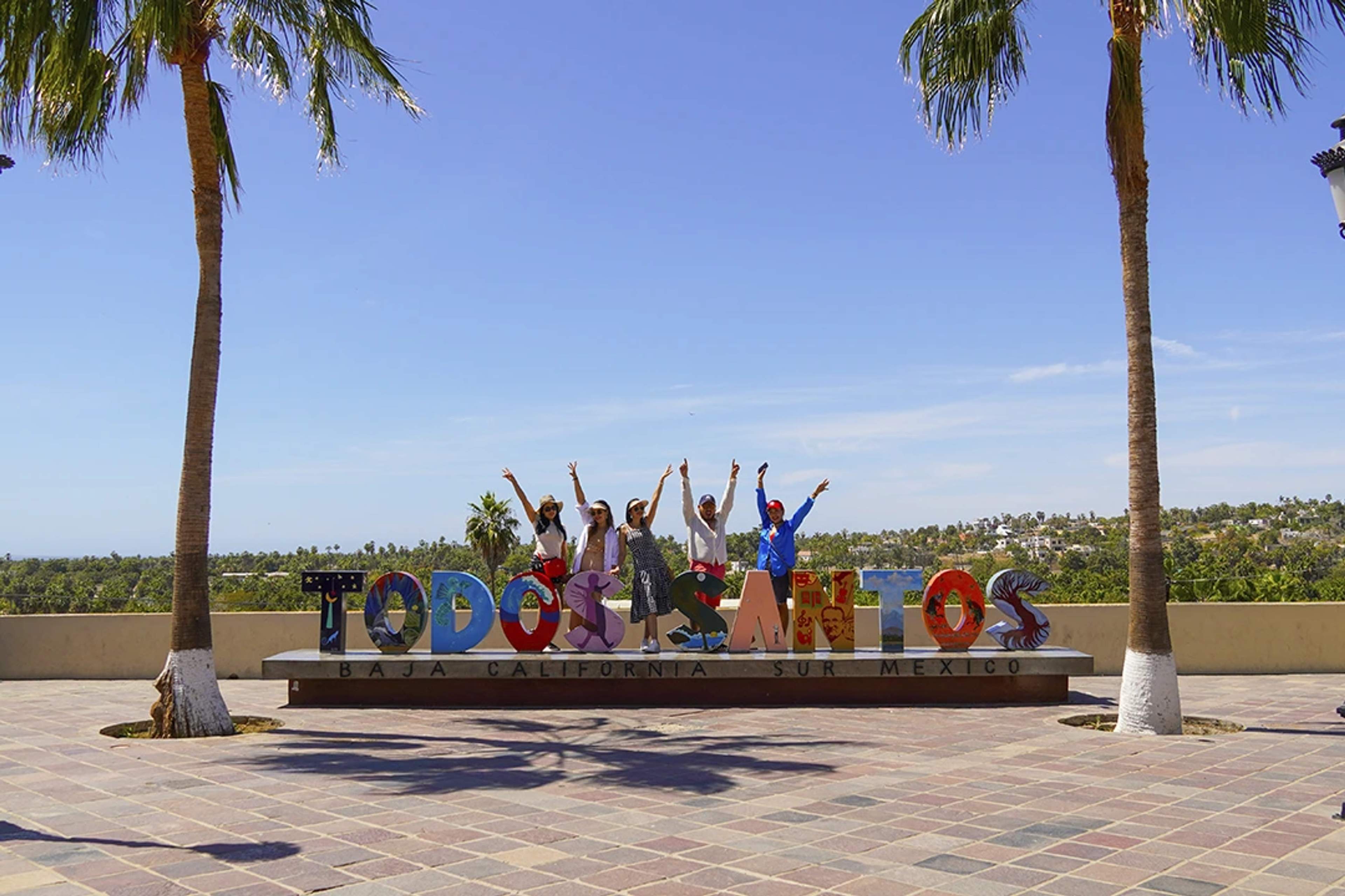 Group of people posing excitedly with the colorful "Todos Santos" sign in Baja California Sur, Mexico, framed by palm trees and blue sky.