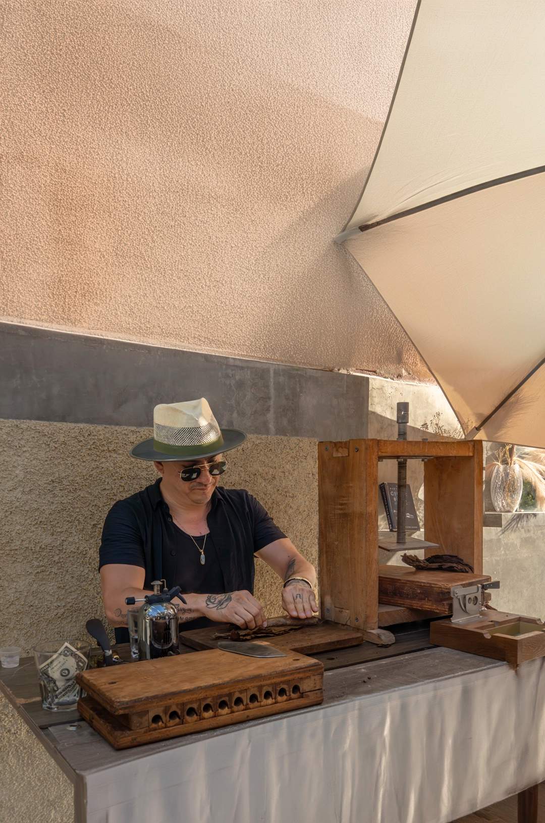 A man wearing a hat and sunglasses rolls a cigar at a wooden table under a shaded area, with cigar-making tools around him.