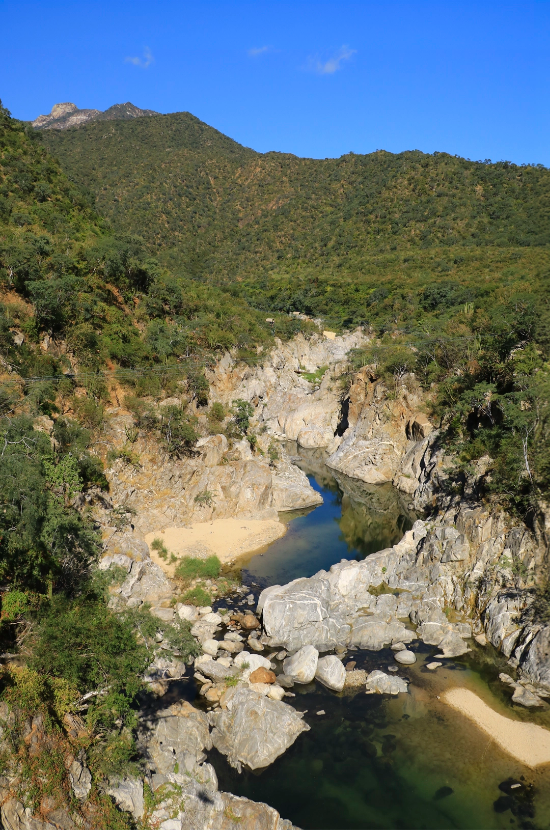 A rocky gorge with a clear stream running through it, surrounded by green hills under a blue sky.