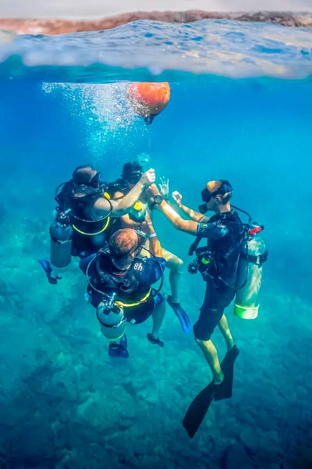 Group of scuba divers practicing safety skills underwater, gathered around a buoy line in clear blue water.