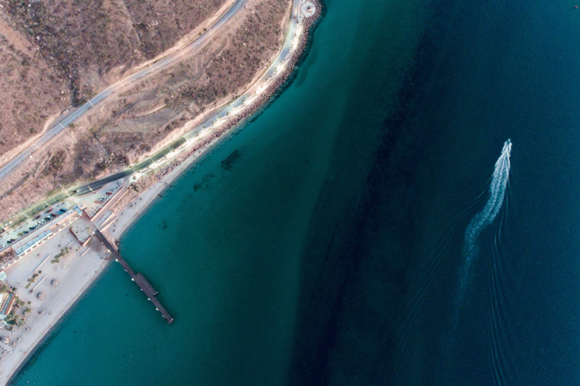 Aerial view of a coastal area in La Paz, Mexico, showing clear blue waters, a pier, a boat leaving a trail, and a rugged shoreline.