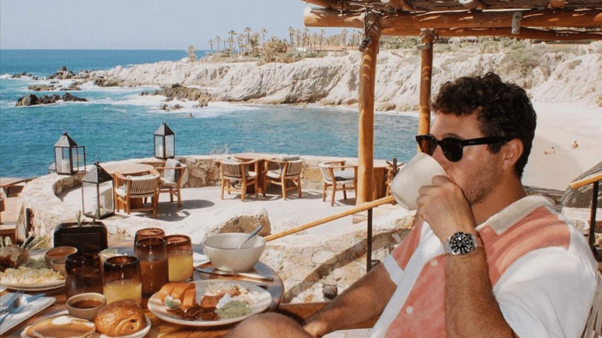 A man enjoying breakfast at an outdoor restaurant overlooking a scenic coastal view, with plates of food and drinks on the table and the ocean in the background.