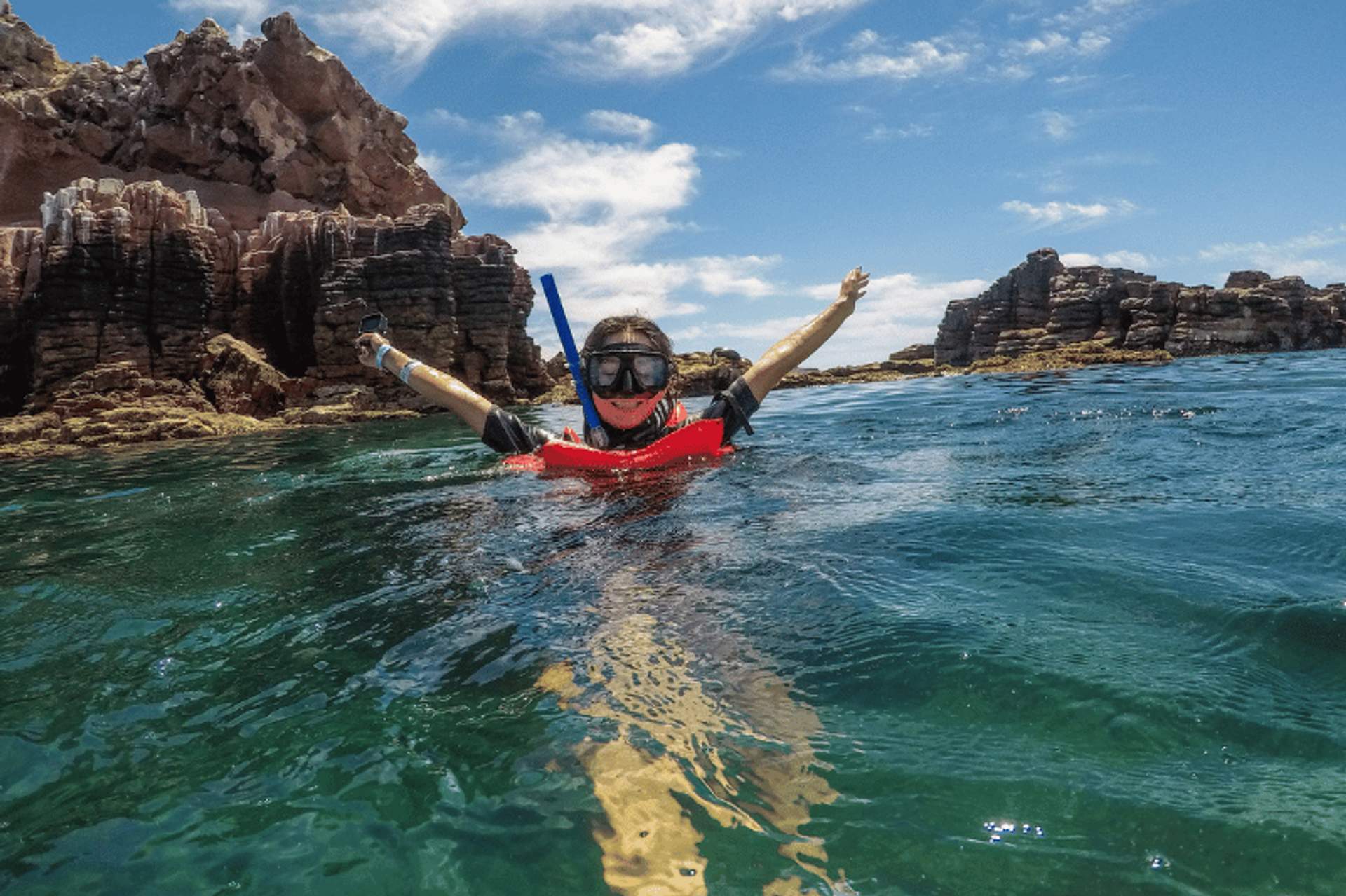 Person snorkeling near rocky cliffs in clear green water, wearing a red life vest and snorkel gear, with arms raised in excitement.