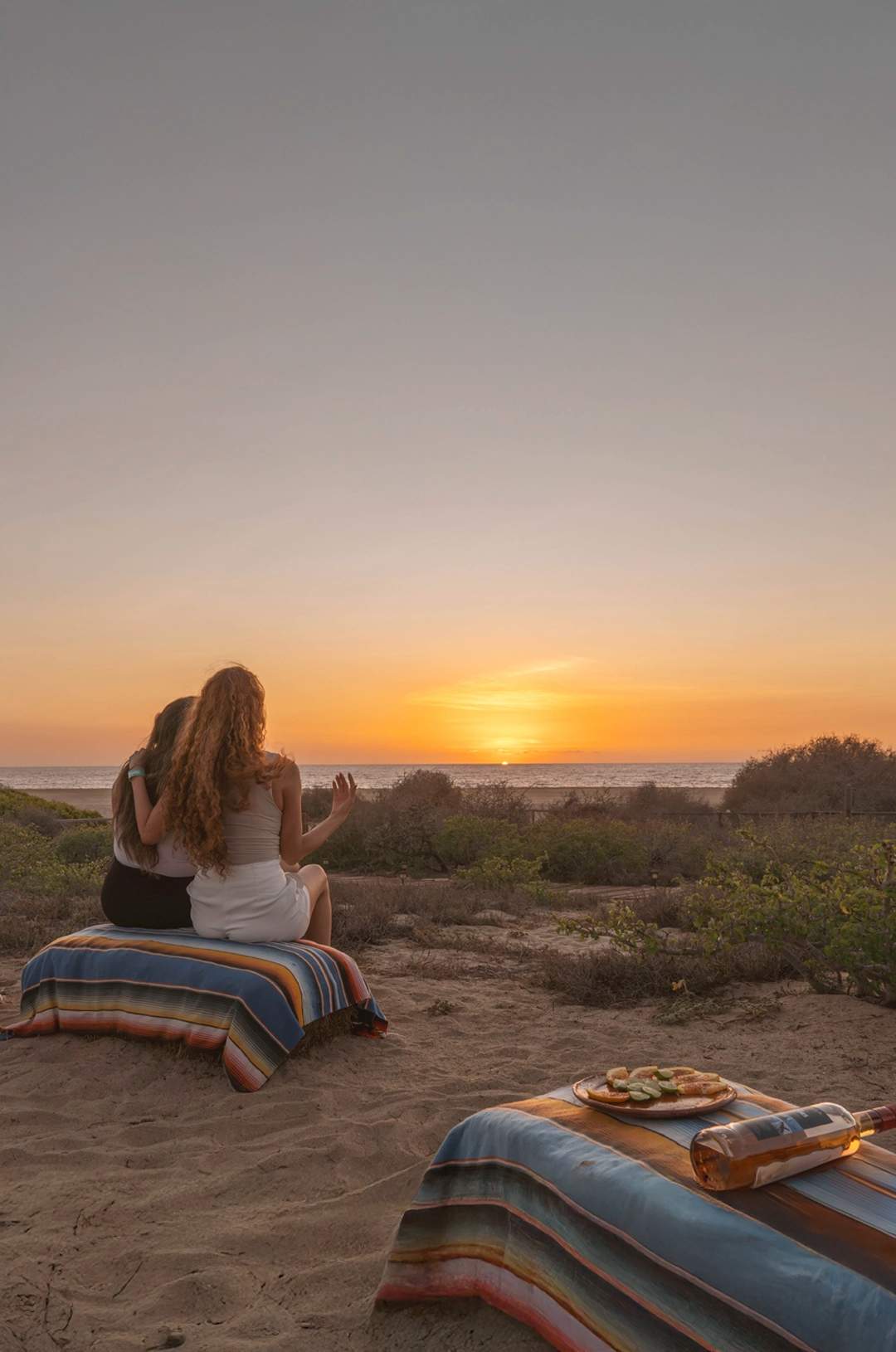 Two people sit on colorful blankets, watching the sunset over the ocean with snacks and drinks nearby on a sandy beach.