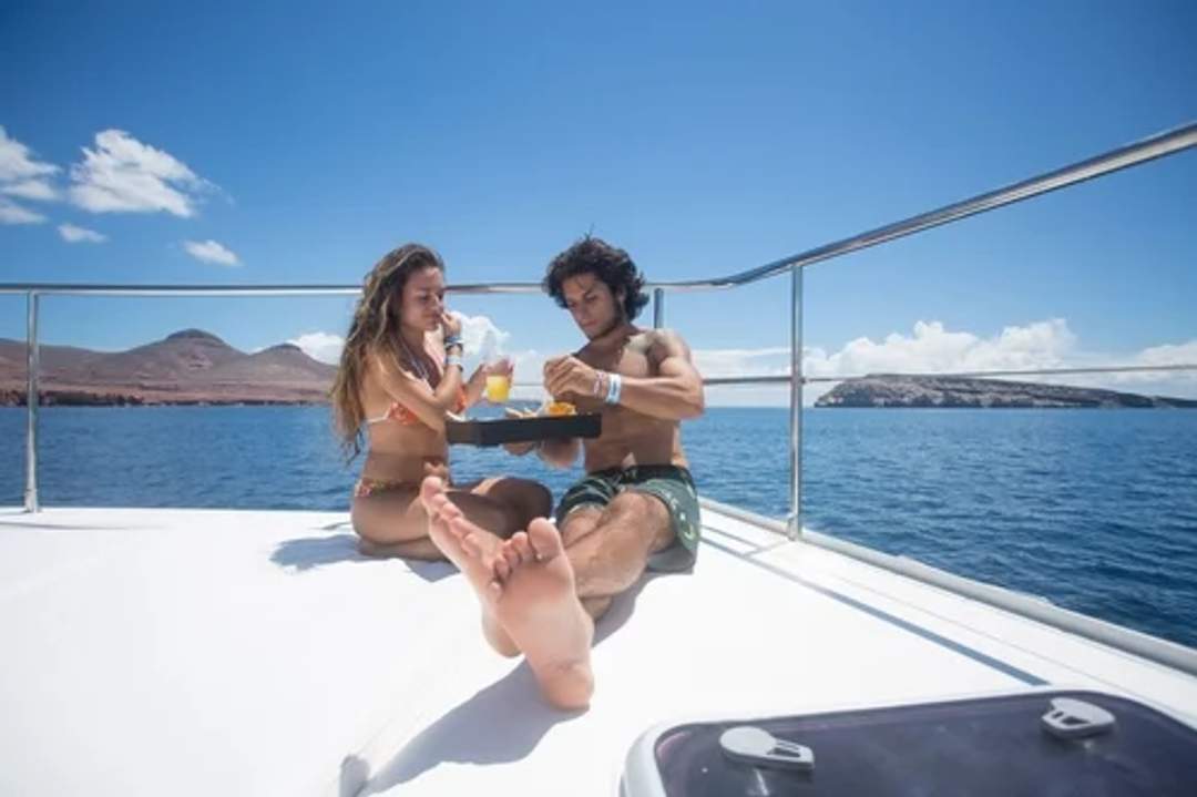 A young couple relaxes and enjoys a luxurious lunch on the bow of a catamaran, with the stunning Cabo coastline as their backdrop.
