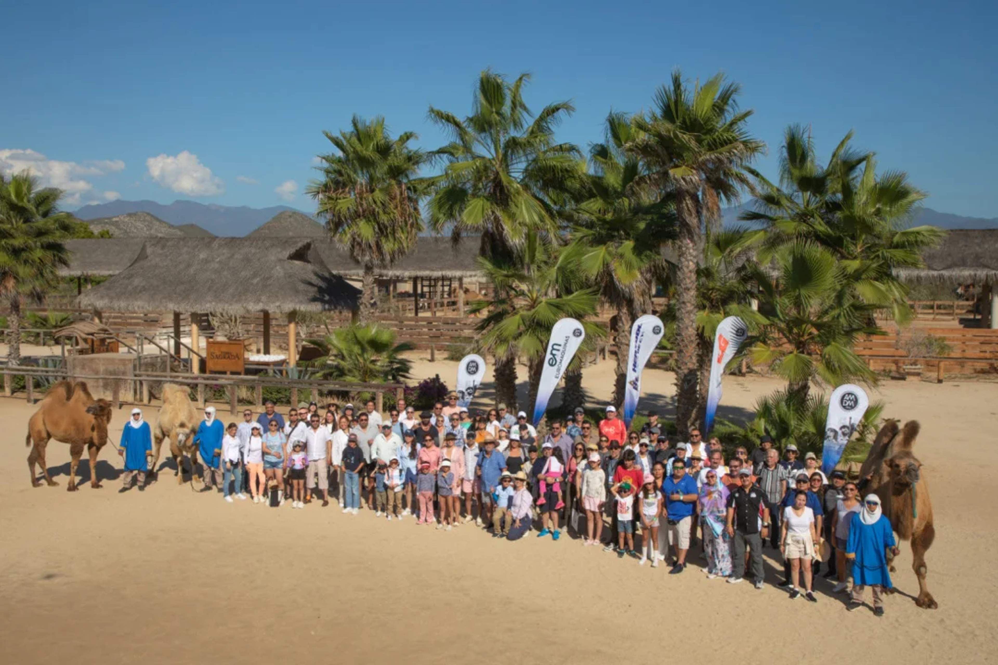 Group of people posing with camels and palm trees at a ranch under a clear sky in Los Cabos.