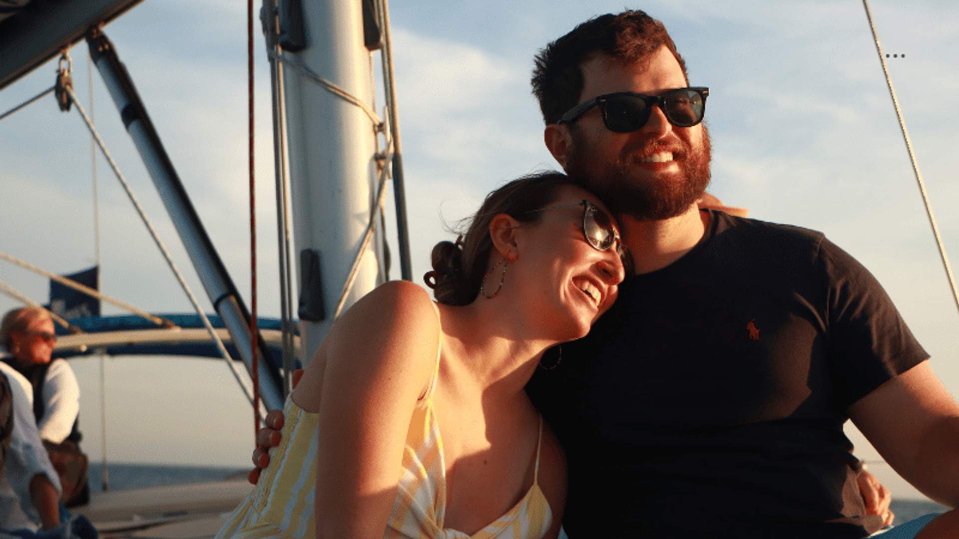 A happy couple enjoys a romantic sunset sailing trip in Cabo, smiling and embracing on the deck of a sailboat.