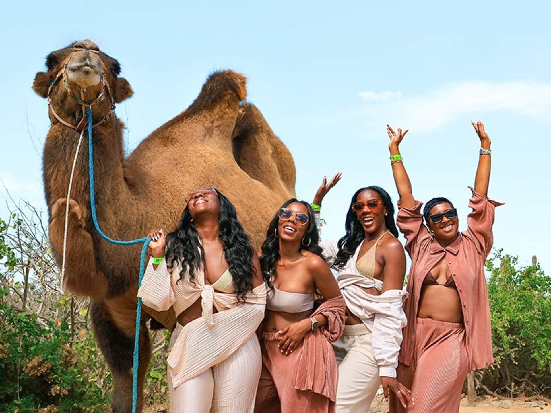 Four women pose and laugh with a camel in a desert setting, enjoying a sunny day.