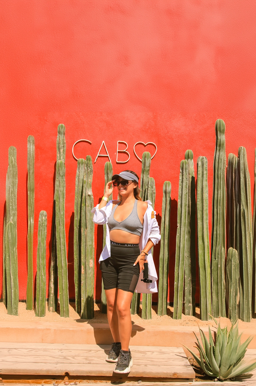 A smiling woman poses in front of a sign that says "Cabo" with cacti and a bright red background.