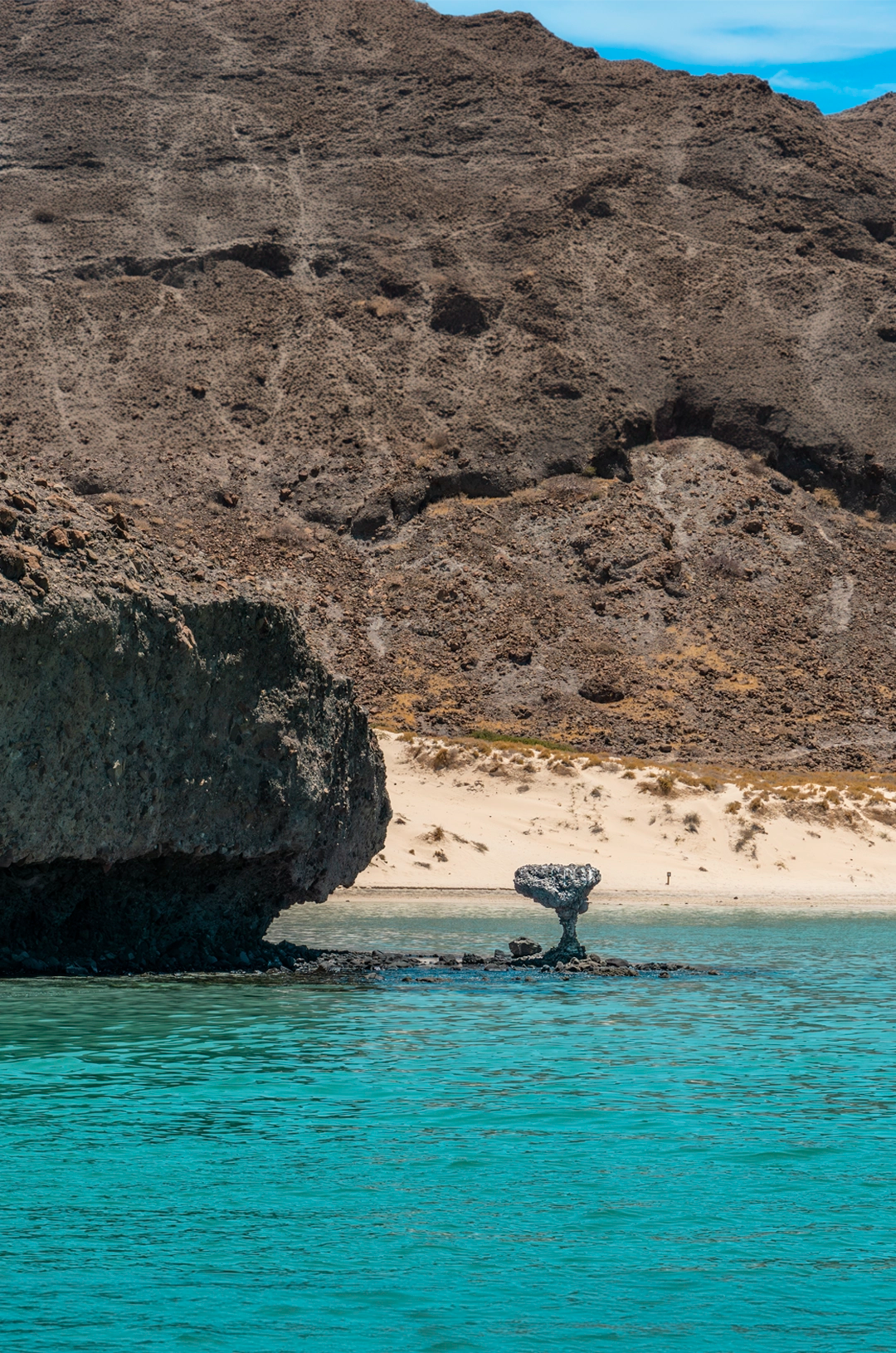 The iconic Balandra Mushroom rock formation in turquoise water with a sandy beach and rocky cliffs in the background.