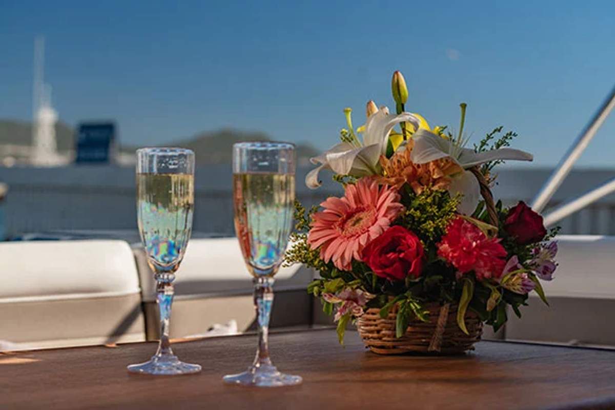 Two champagne flutes sit beside a colorful flower arrangement on a wooden table aboard a yacht. The elegant setup against a backdrop of calm waters and distant hills suggests a celebration or romantic occasion at sea.