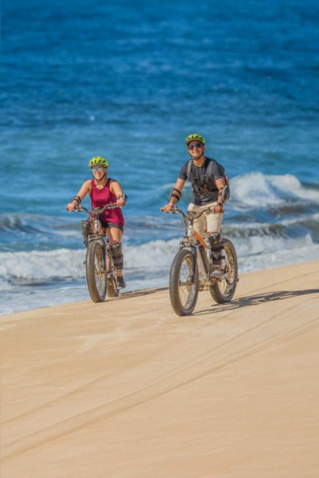 Electric bike tour in Cabo, couple riding along a sandy beach with ocean waves in the background.