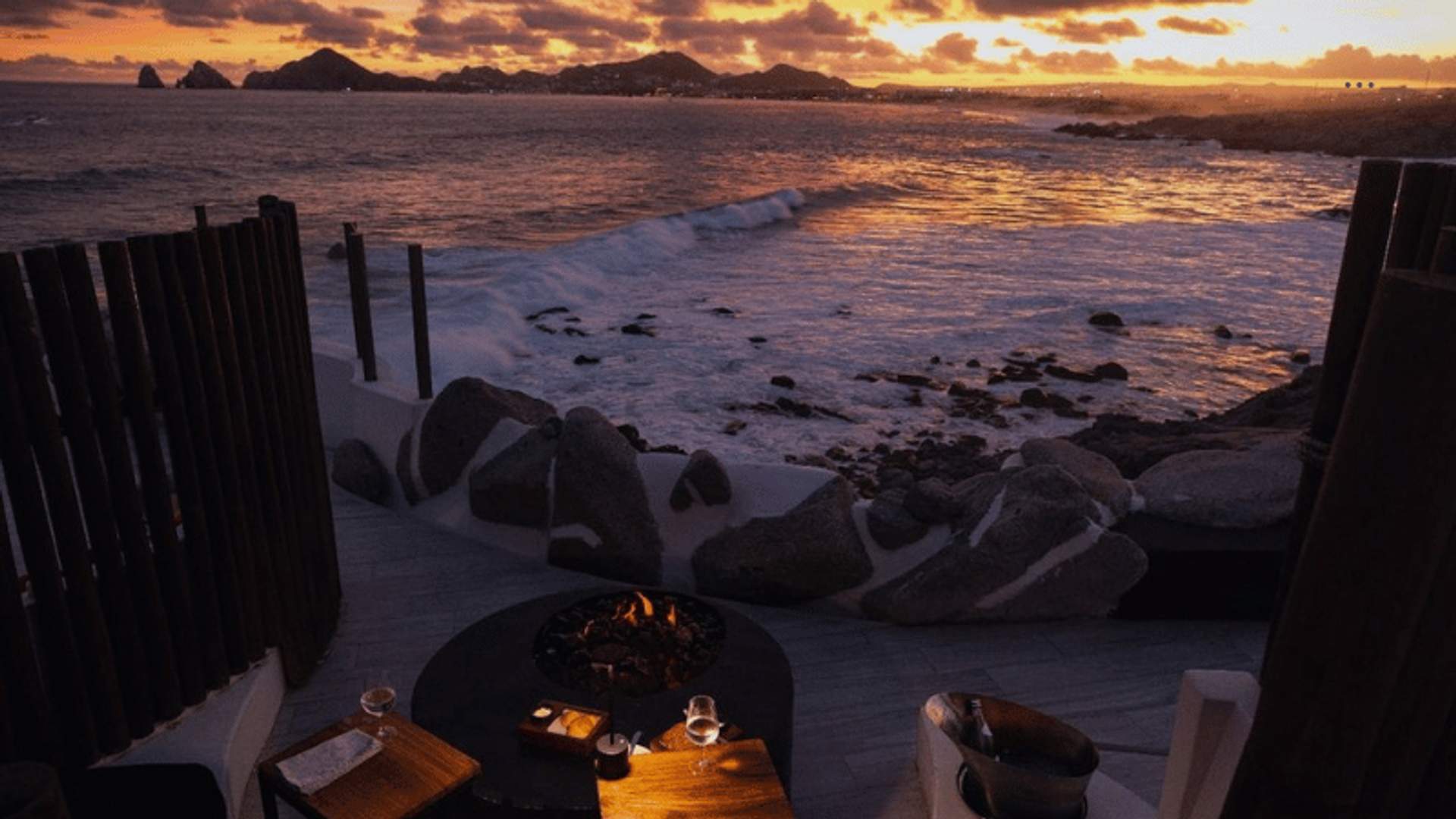 Sunset view from an outdoor restaurant by the ocean, featuring a cozy seating area with a lit fire pit and a table set for two.