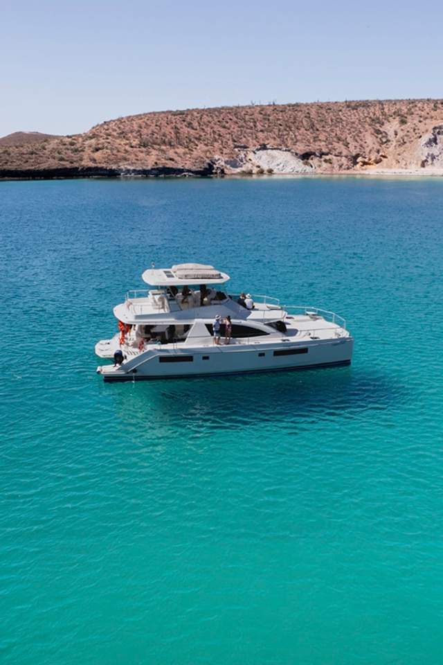 A white catamaran sailing on clear turquoise waters near a rocky coastline with a few people on board.