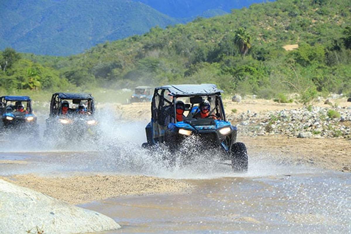 A group of off-road vehicles drives through a shallow stream on a rugged trail, creating splashes and waves as they traverse the water. The scene is set against a backdrop of green hills and blue mountains under a clear sky.