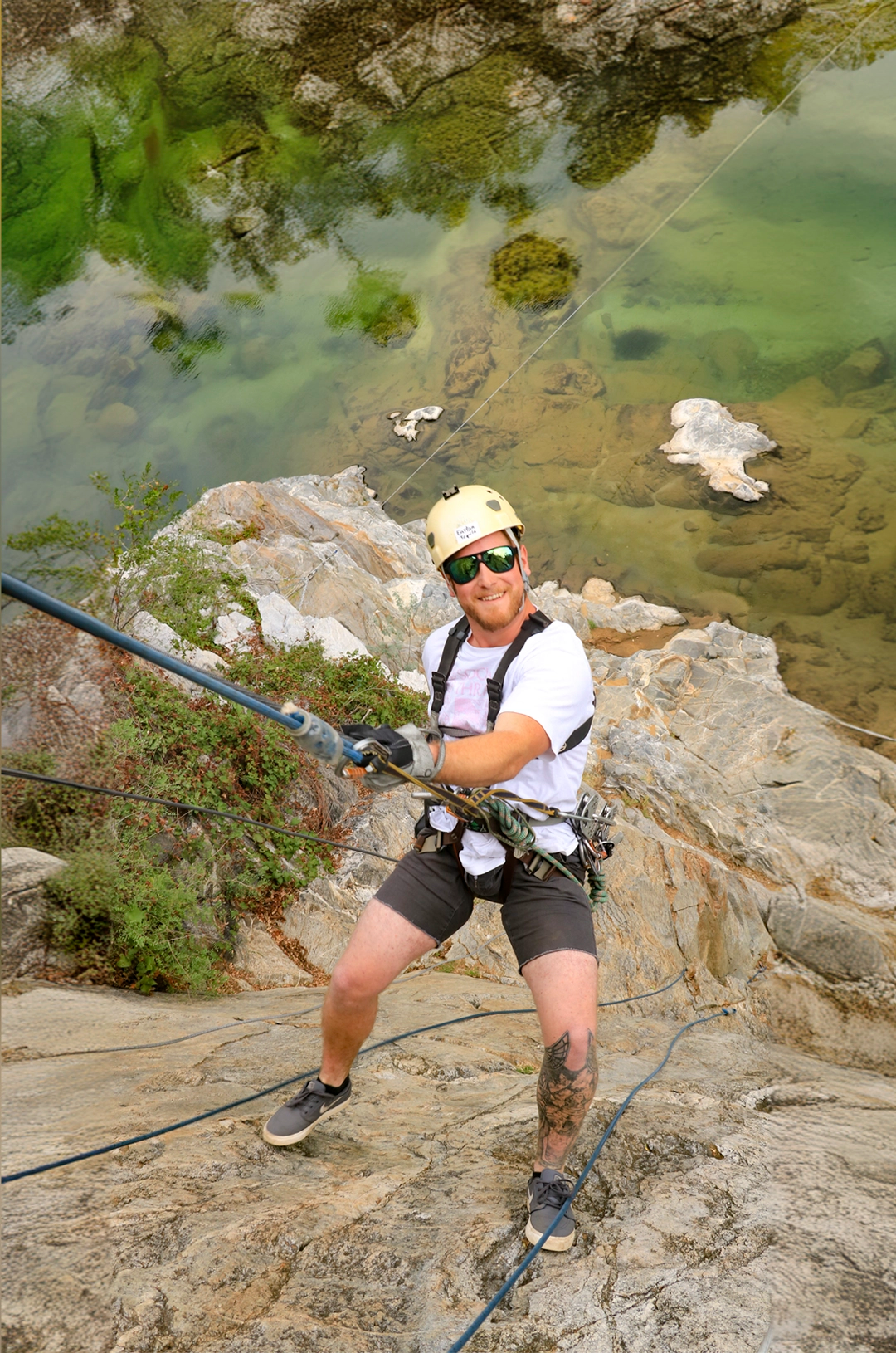 A man rappelling down a rock face above a clear green water pool, wearing a helmet, sunglasses, and harness.