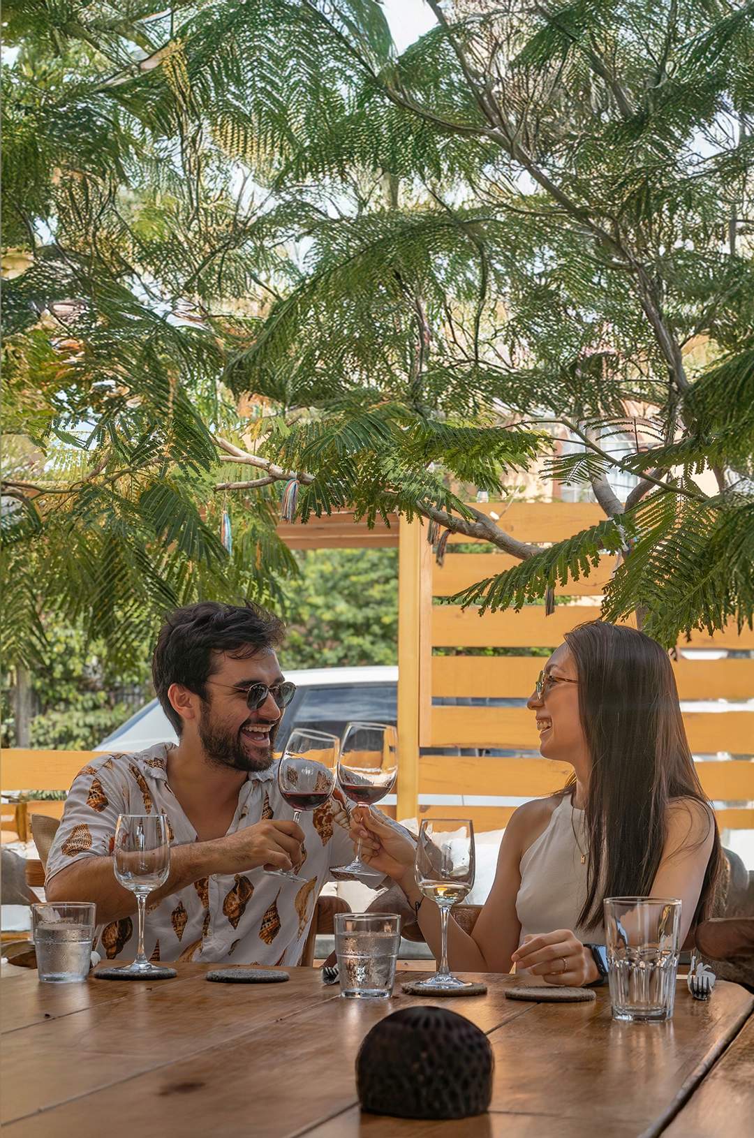 Smiling couple toasting with wine at an outdoor table, surrounded by greenery in a relaxed setting.