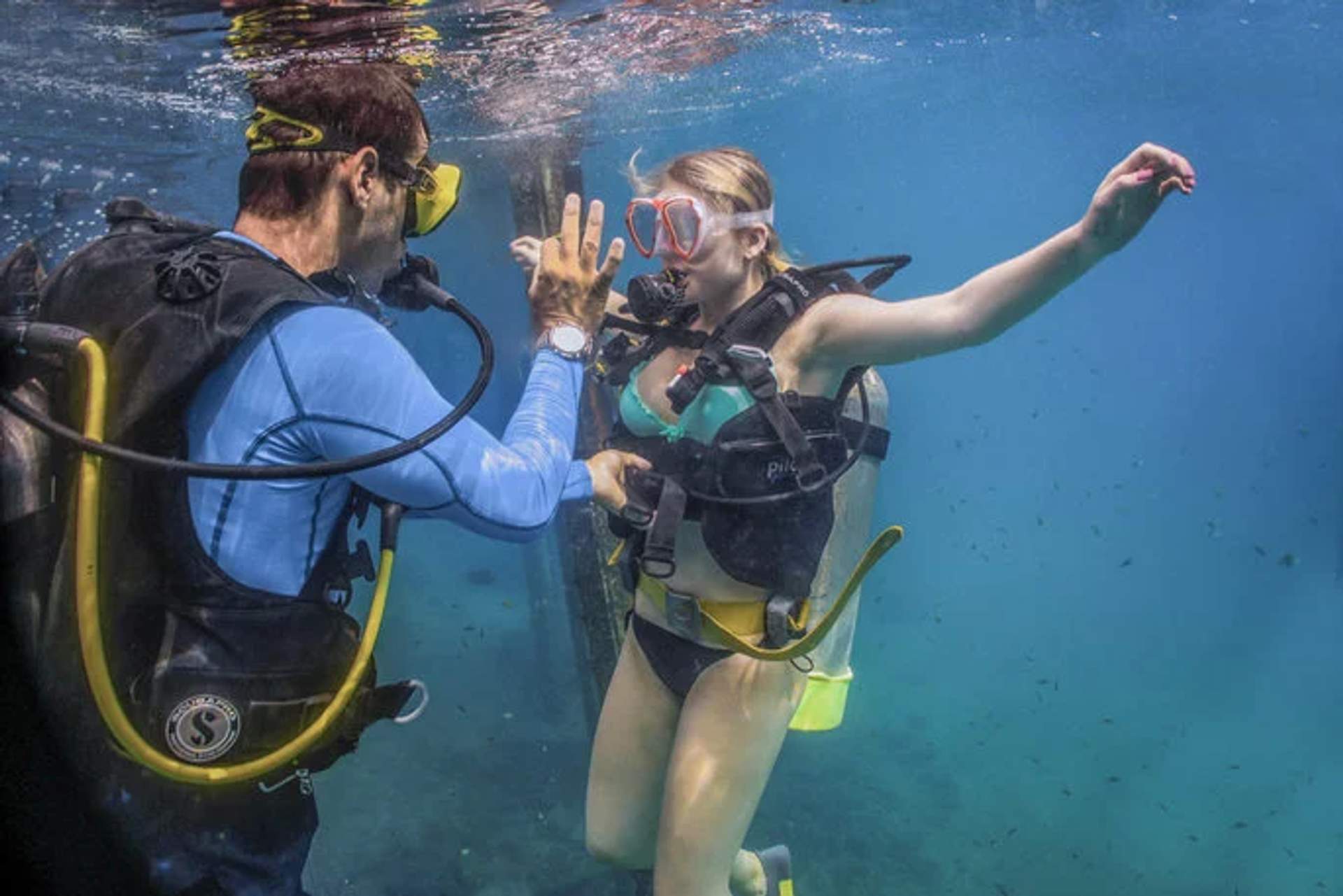 A scuba diving instructor guiding a student underwater, both equipped with diving gear. The instructor is giving a hand signal, possibly for communication or instruction, while the student attentively listens and maintains buoyancy. The underwater setting is clear, with various aquatic elements visible in the background.