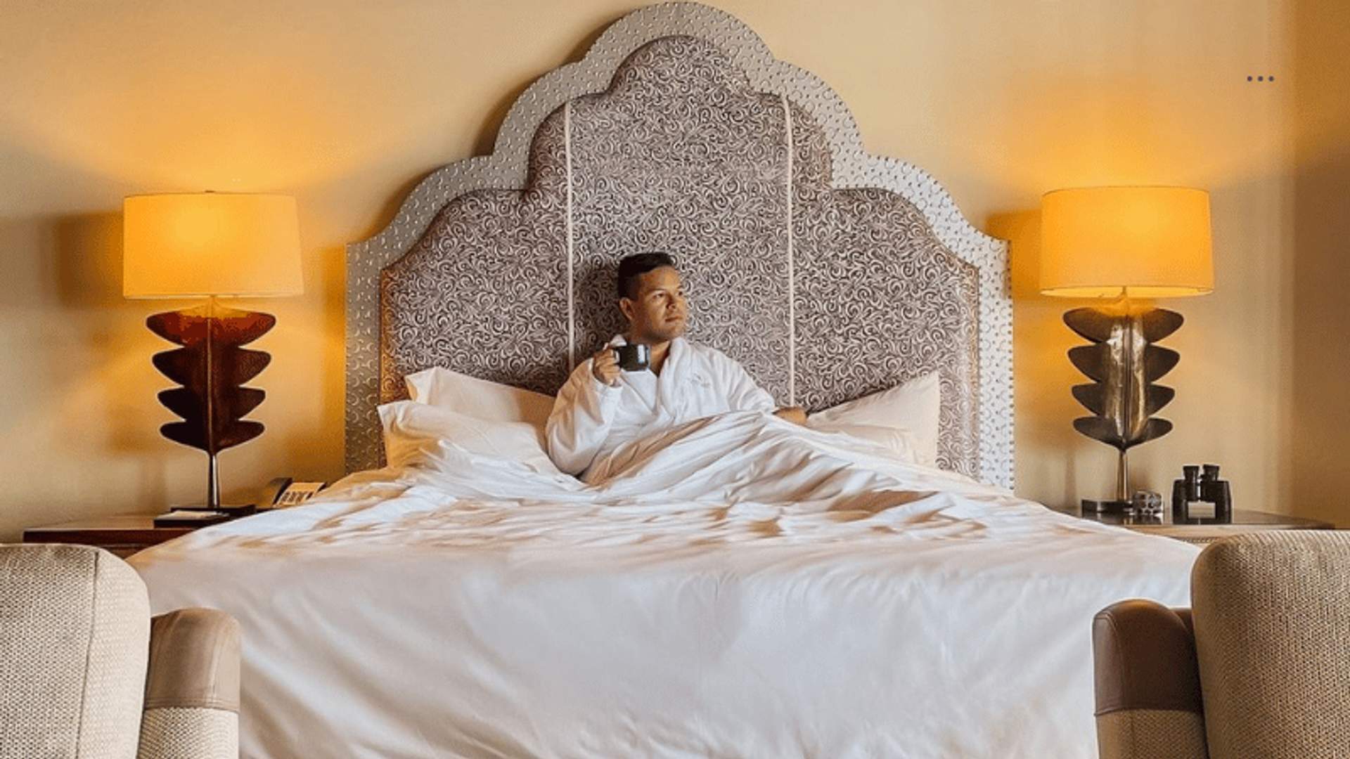 A man sitting in a luxurious hotel bed, wearing a white bathrobe and holding a cup of coffee, with two bedside lamps casting a warm glow.