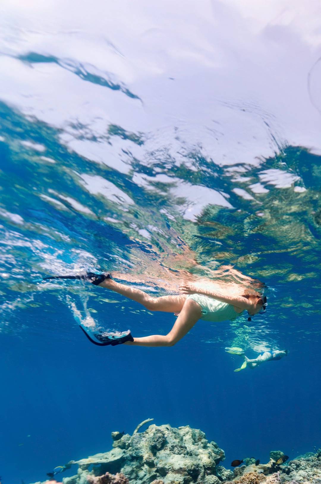Woman snorkeling above a coral reef, surrounded by clear blue water, with fish visible near the seafloor.