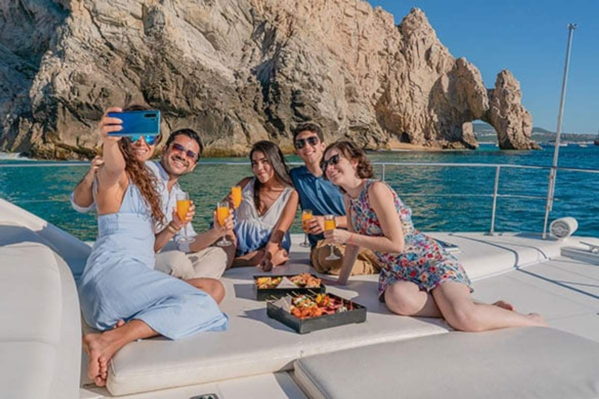 A group of five friends are on a yacht, posing for a selfie while holding orange drinks. They are enjoying a sunny day with a scenic backdrop of rocky cliffs and a natural arch formation over the ocean. There is a tray of food on the deck beside them.