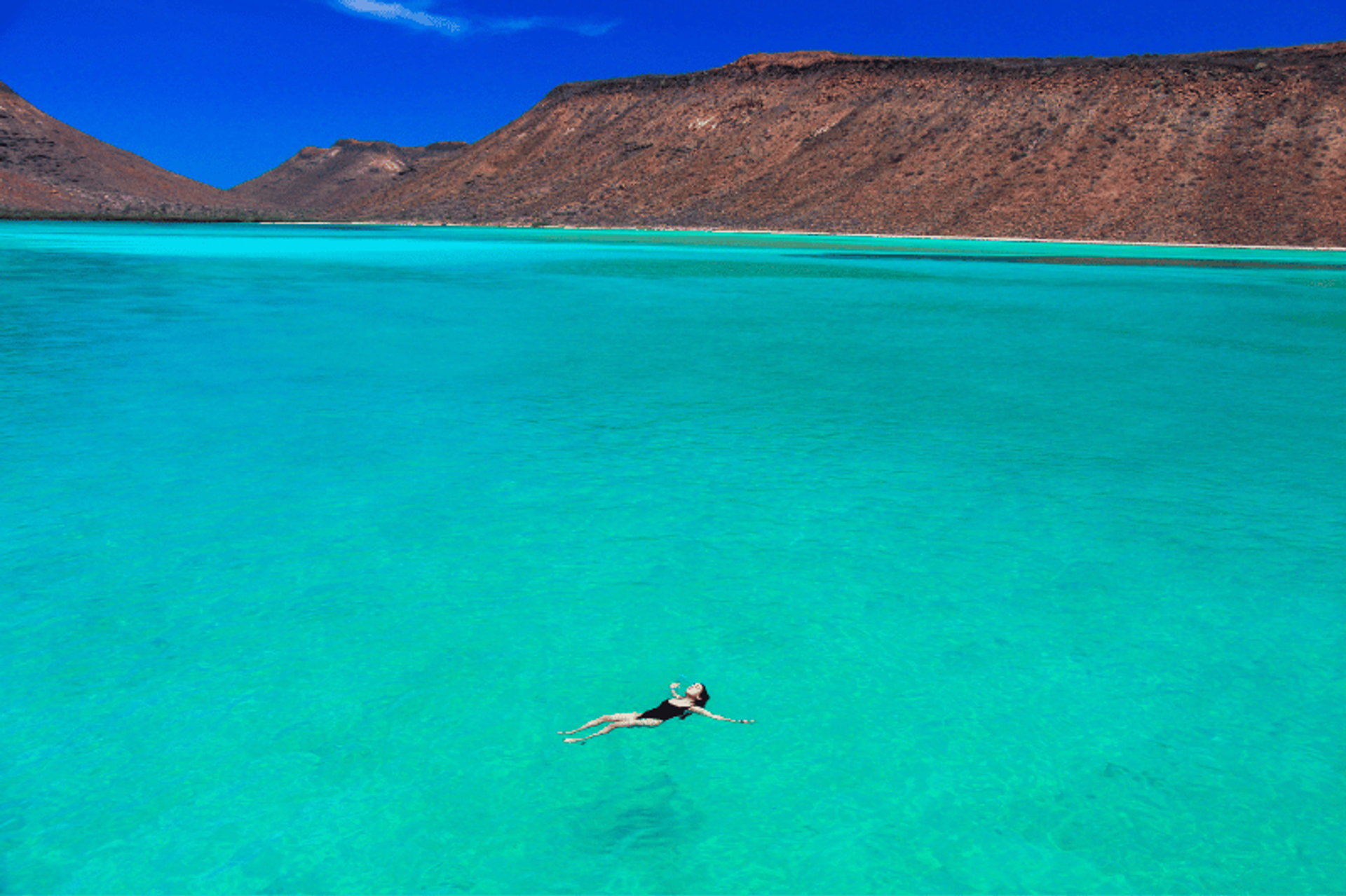 Person floating in crystal-clear turquoise water with rugged, desert mountains in the background under a bright blue sky.