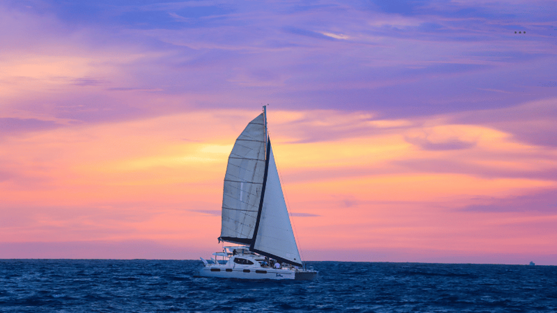 A sailboat cruising on the ocean at sunset, with a vibrant sky painted in shades of purple, pink, and orange.