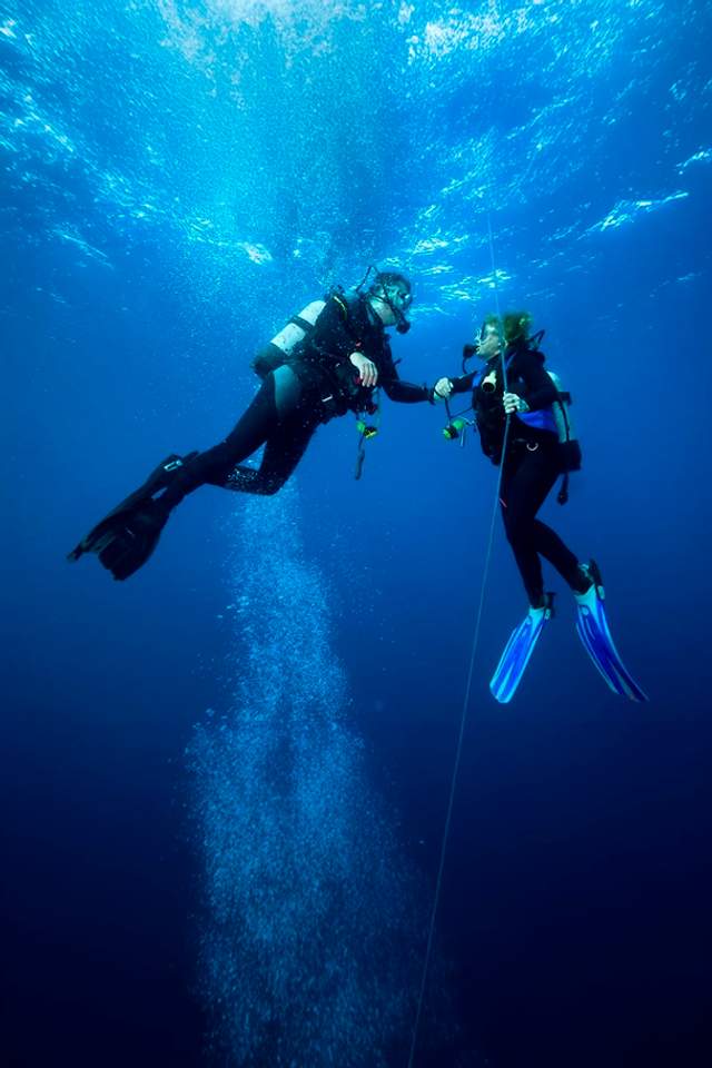 Two scuba divers hold onto a guide rope underwater, communicating as bubbles rise around them in the deep blue ocean.