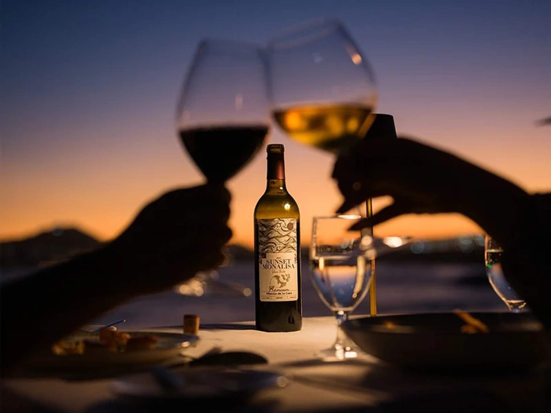 Two people toasting with wine glasses at sunset, with a bottle of Sunset Monalisa wine on the table.