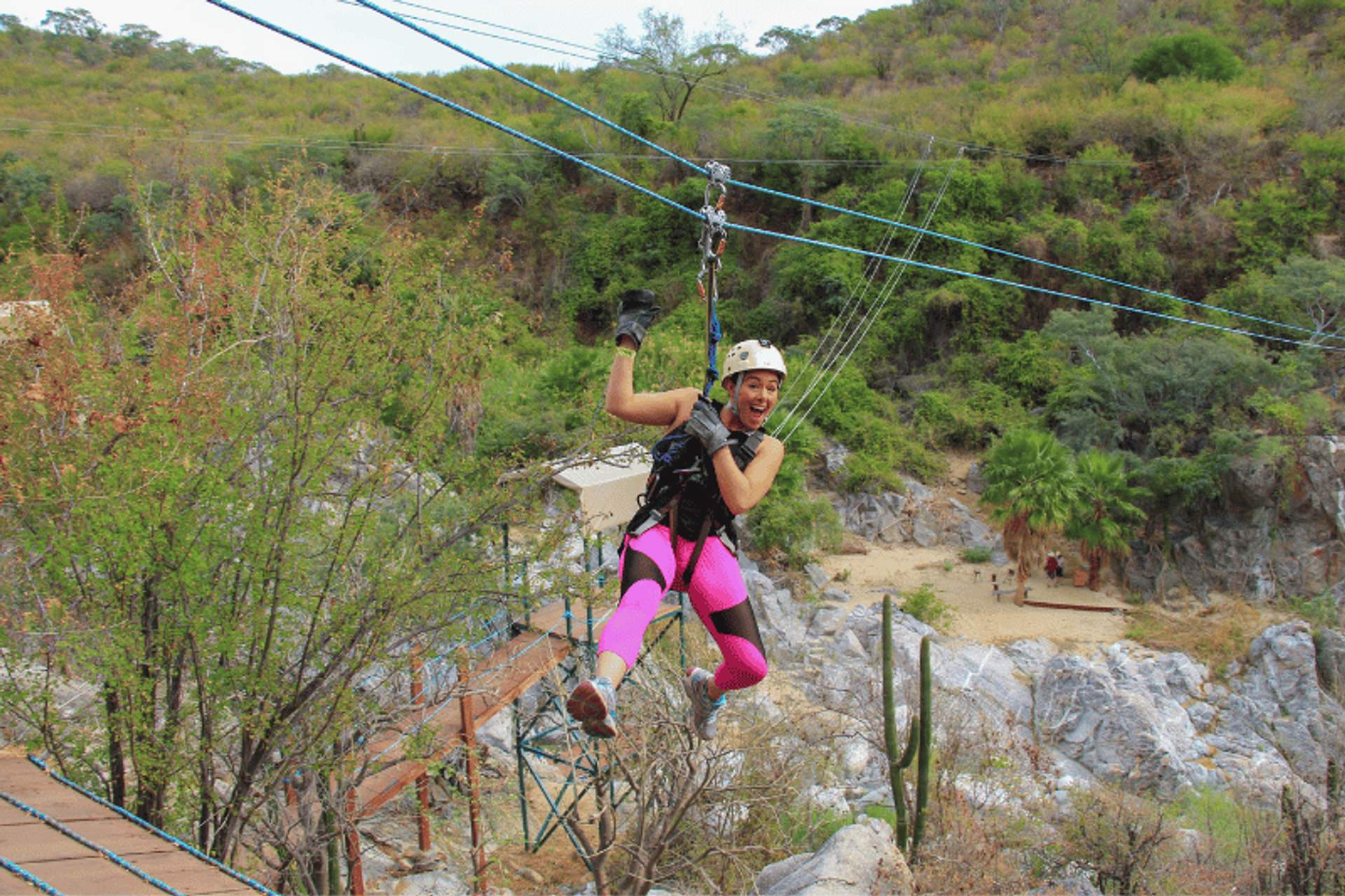 A person wearing a helmet and bright pink leggings enjoying a zipline ride through a lush, green landscape with trees and rocks in the background.