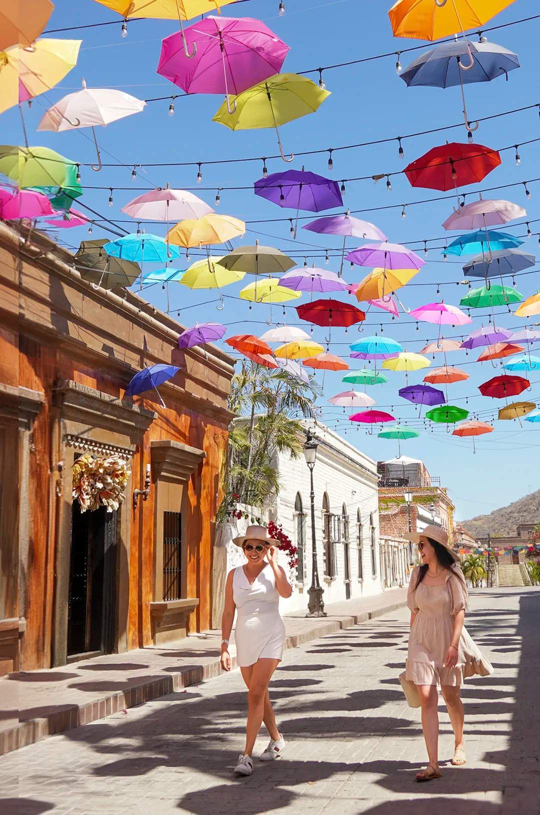 Two women walking on a sunny street with colorful umbrellas hanging above, wearing summer hats and dresses, and smiling.
