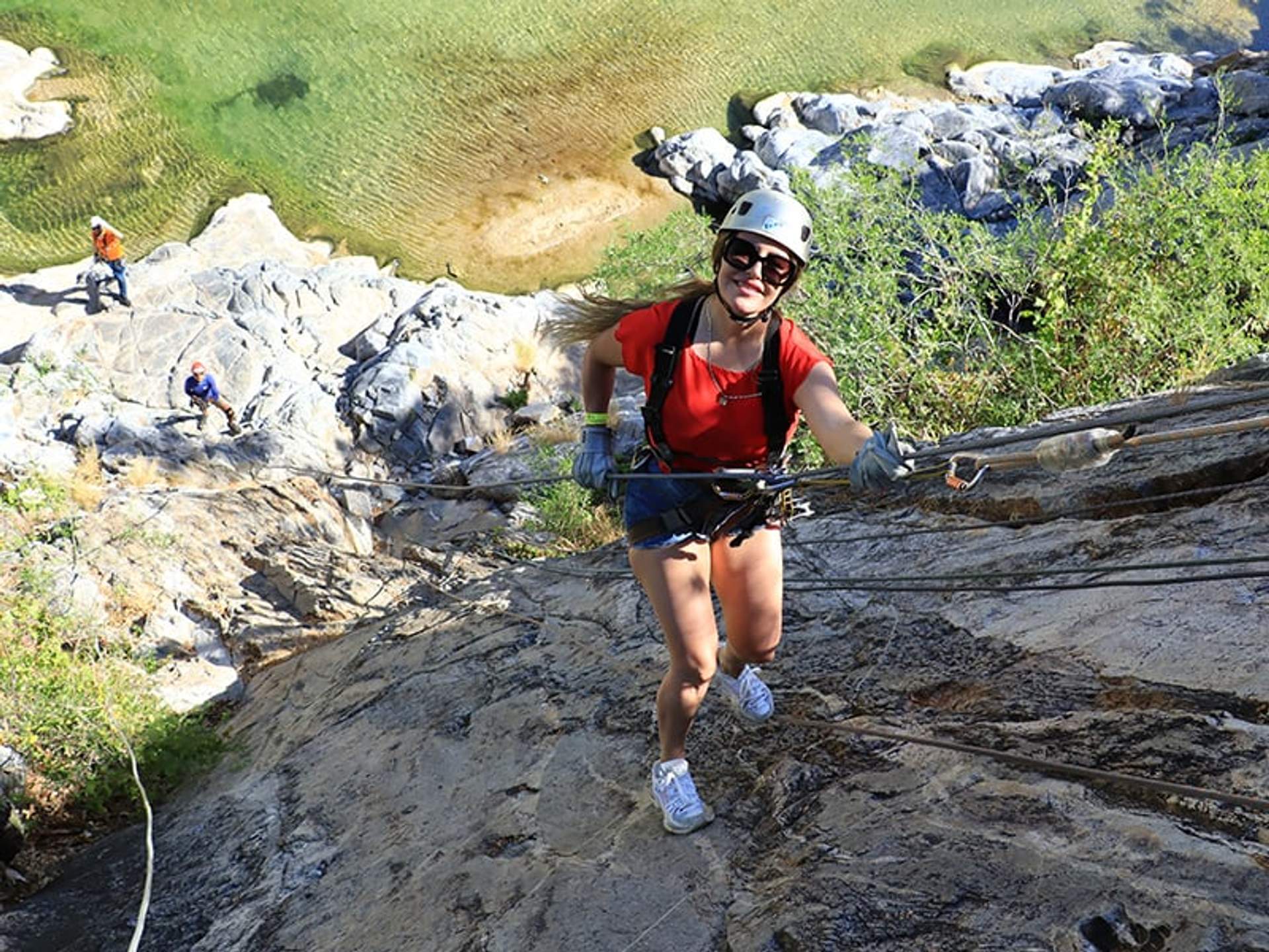 A woman in a helmet and harness smiles while rappelling down a rocky cliff, with a river and other climbers below.