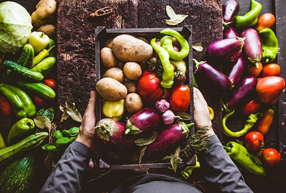 A person holding a crate of fresh vegetables, including potatoes, tomatoes, eggplants, and peppers, surrounded by more vegetables on a rustic table.