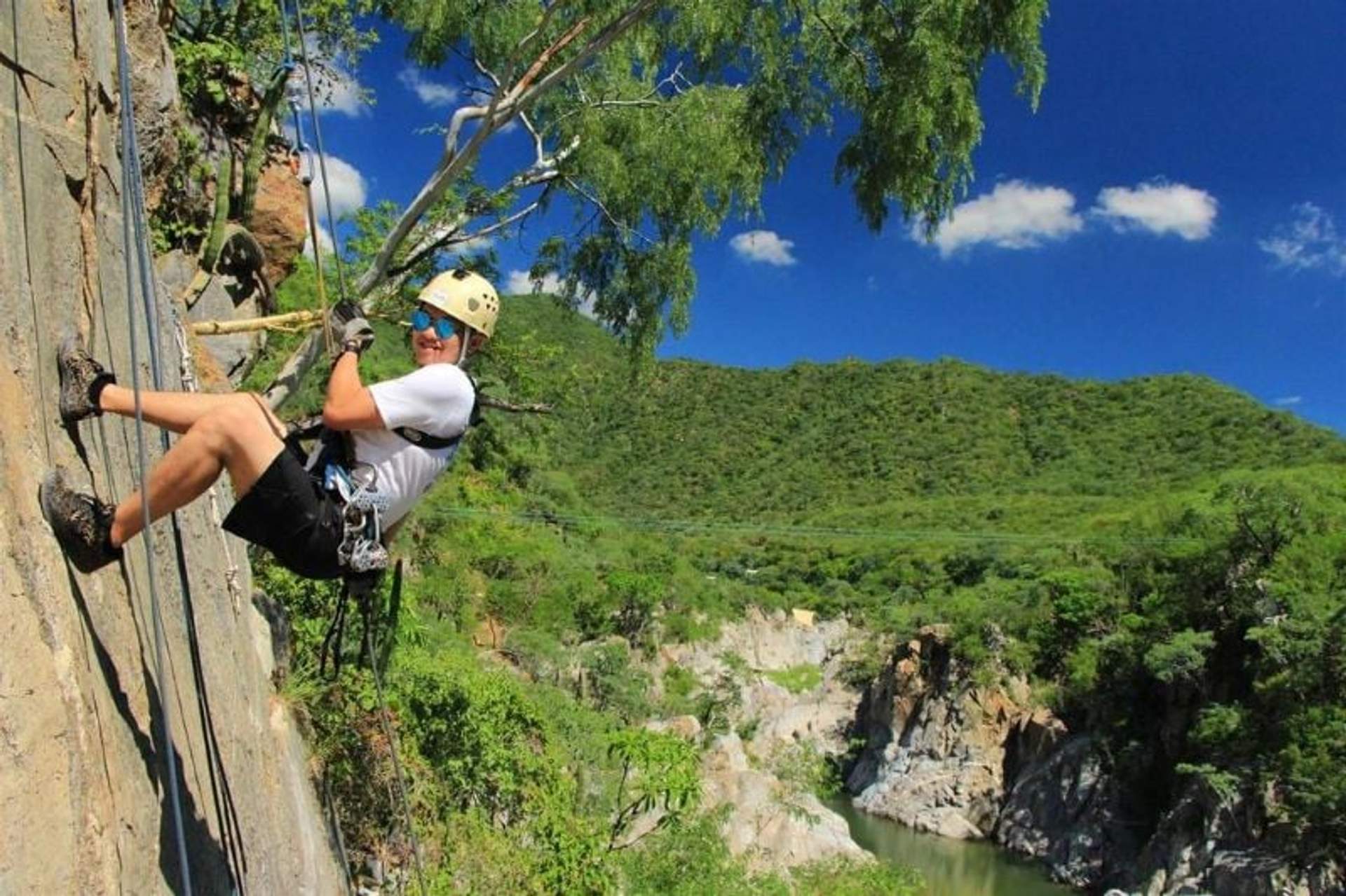Person rappelling down a rock face with a lush green valley and river in the background, smiling and wearing safety gear.