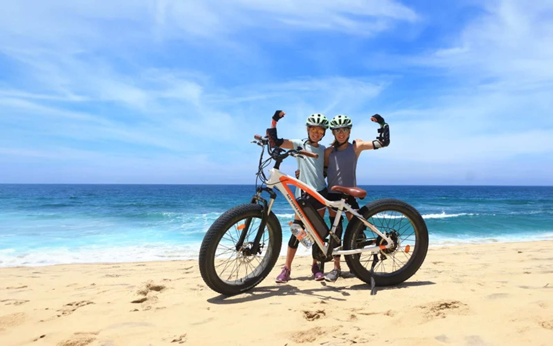 Two cyclists on a beach, wearing helmets and protective gear, pose next to their bicycles under a clear sky.