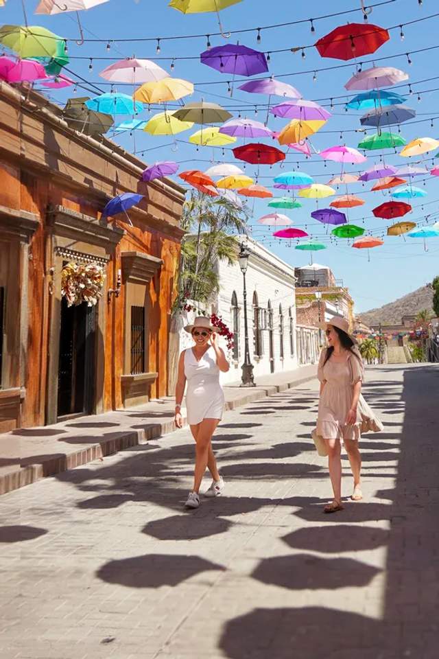 Group jumping in front of the colorful "Todos Santos" sign in Baja California Sur, Mexico, with palm trees and a blue sky.