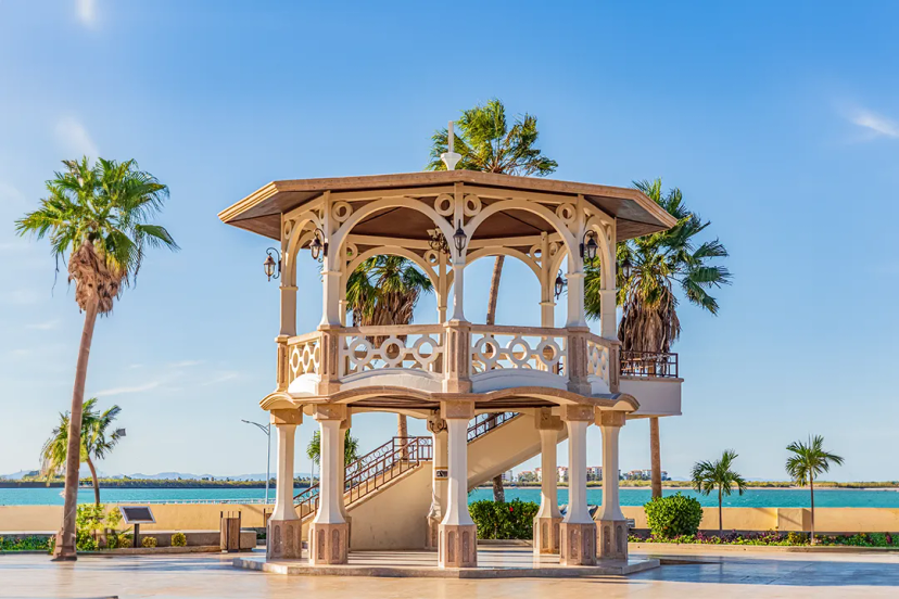 A picturesque gazebo with intricate designs stands near palm trees by the water, under a clear blue sky.