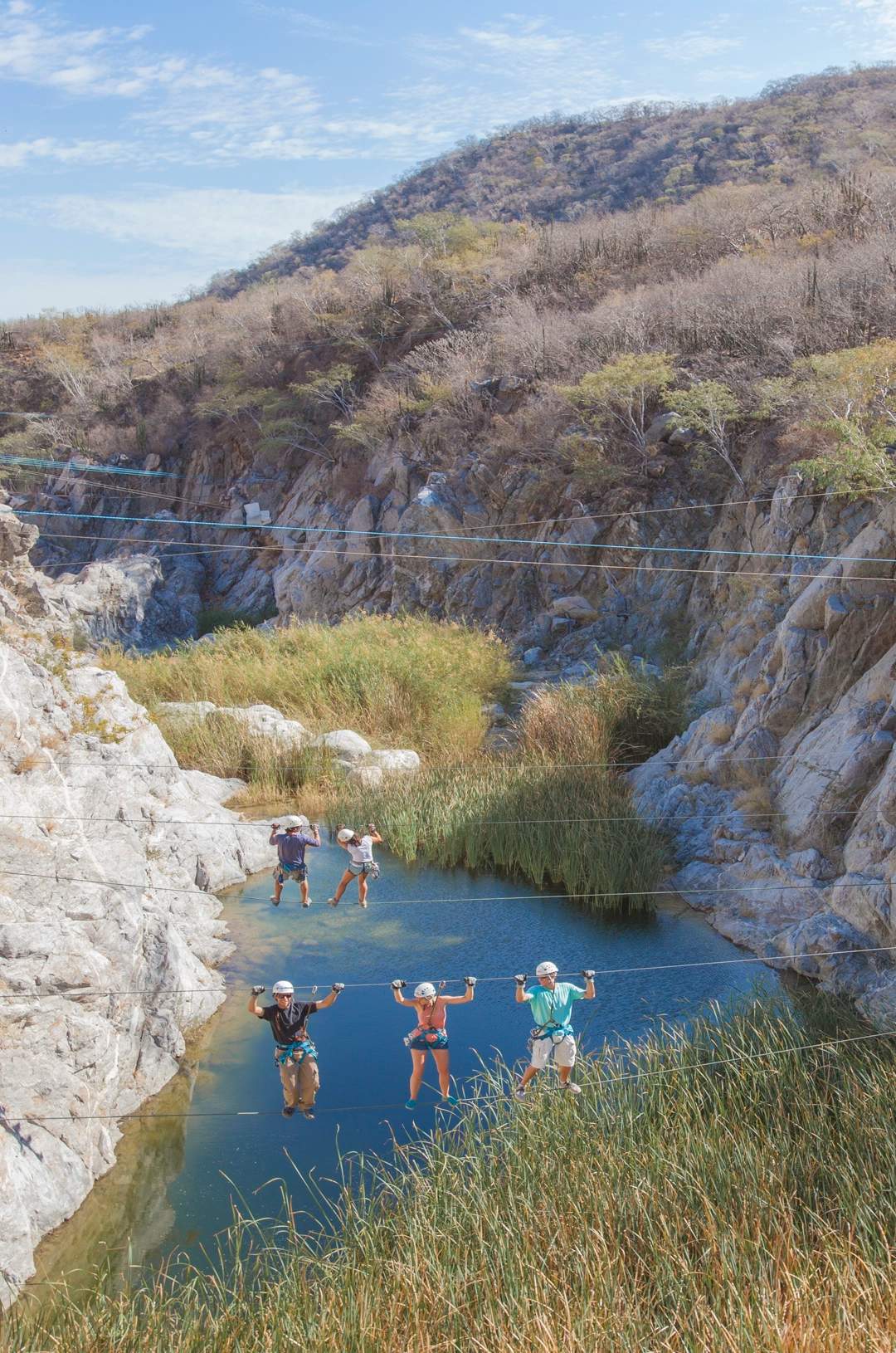 People crossing a narrow suspension bridge over a gorge with a small pool below, wearing helmets and harnesses.