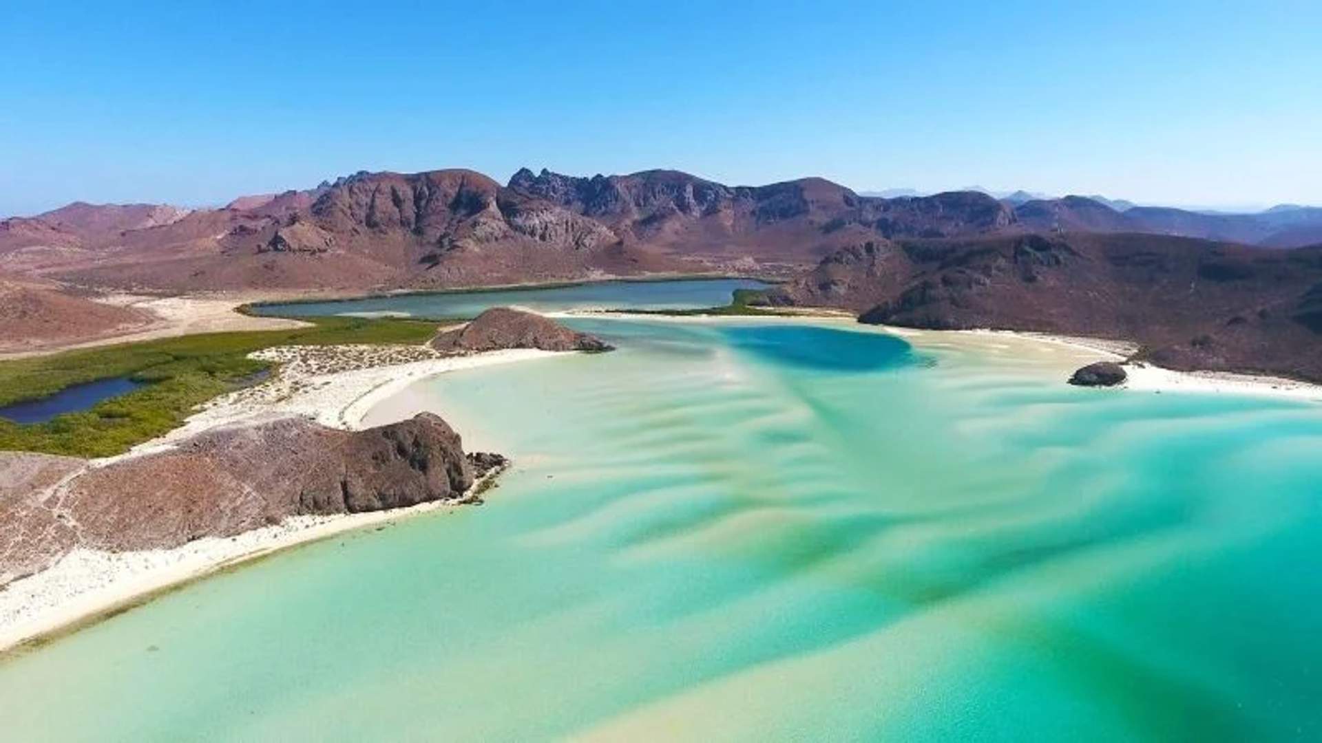 Aerial view of Balandra Beach with turquoise waters, white sandy shores, and mountainous landscape in the background.