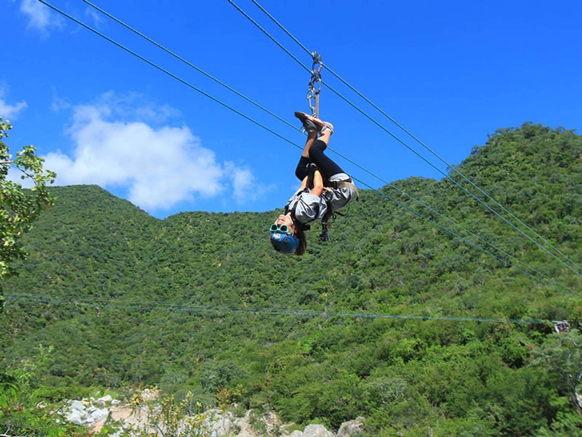A person wearing a helmet and harness hangs upside down while ziplining over a lush, green landscape.