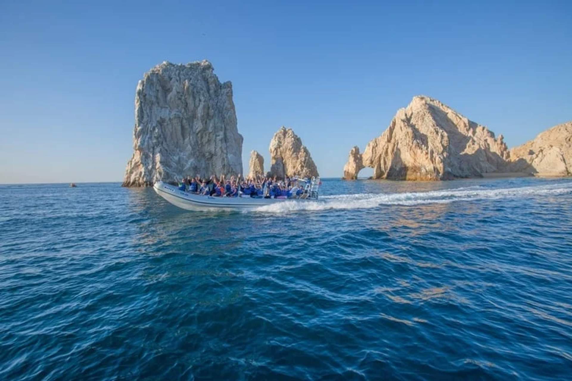 A boat filled with people speeds through the ocean, passing by the iconic rock formations of El Arco at Cabo San Lucas under a clear blue sky. The rugged cliffs and archways rise dramatically from the water, creating a picturesque scene.