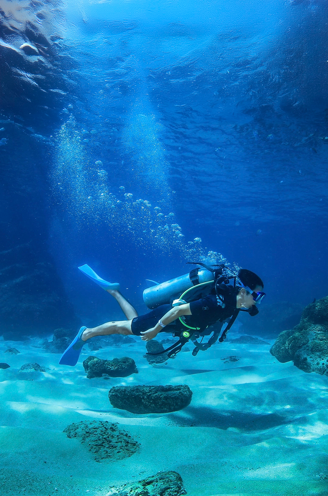 A scuba diver swims over a sandy ocean floor, surrounded by rocks, with bubbles rising in the clear blue water.