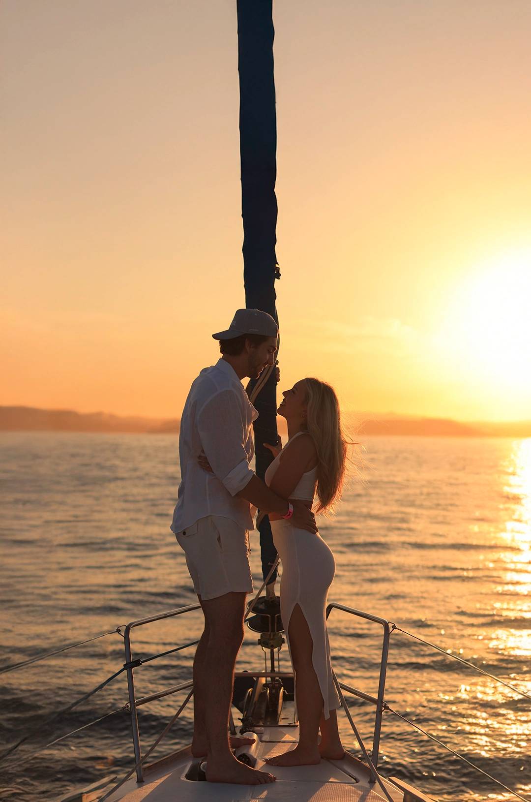 A couple shares a romantic moment on a sailboat at sunset, with golden light reflecting off the ocean.
