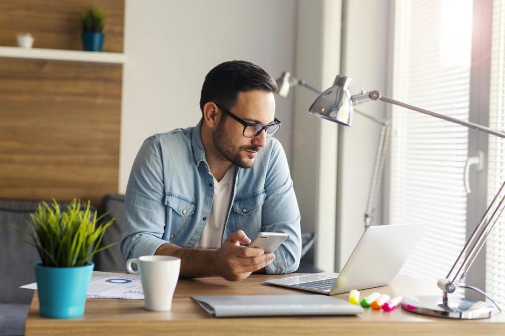 Man wearing collared shirt sitting at desk holding phone and looking at computer